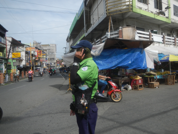 Ein Polizeibeamter in grüner und blauer Uniform und blauer Mütze steht an der Seite einer Straße und spricht auf einem Handy, mit Fahrzeugen, die hinter ihm auf der Straße fahren, Ständen mit verschiedenen Gegenständen rechts von ihm und einem bewölkten Himmel mit einem Gebäude mit Fenstern im Hintergrund.