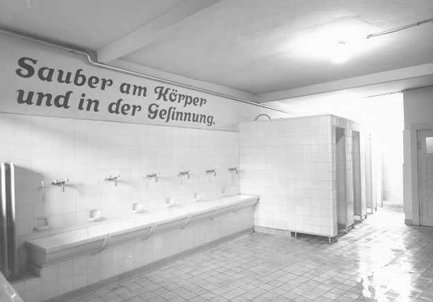 Schwarz-weiß-Foto einer öffentlichen Toilette mit Fliesenboden, einer Wand mit Text, mehreren Wasserhähnen, einer Tür und einer Deckenleuchte.