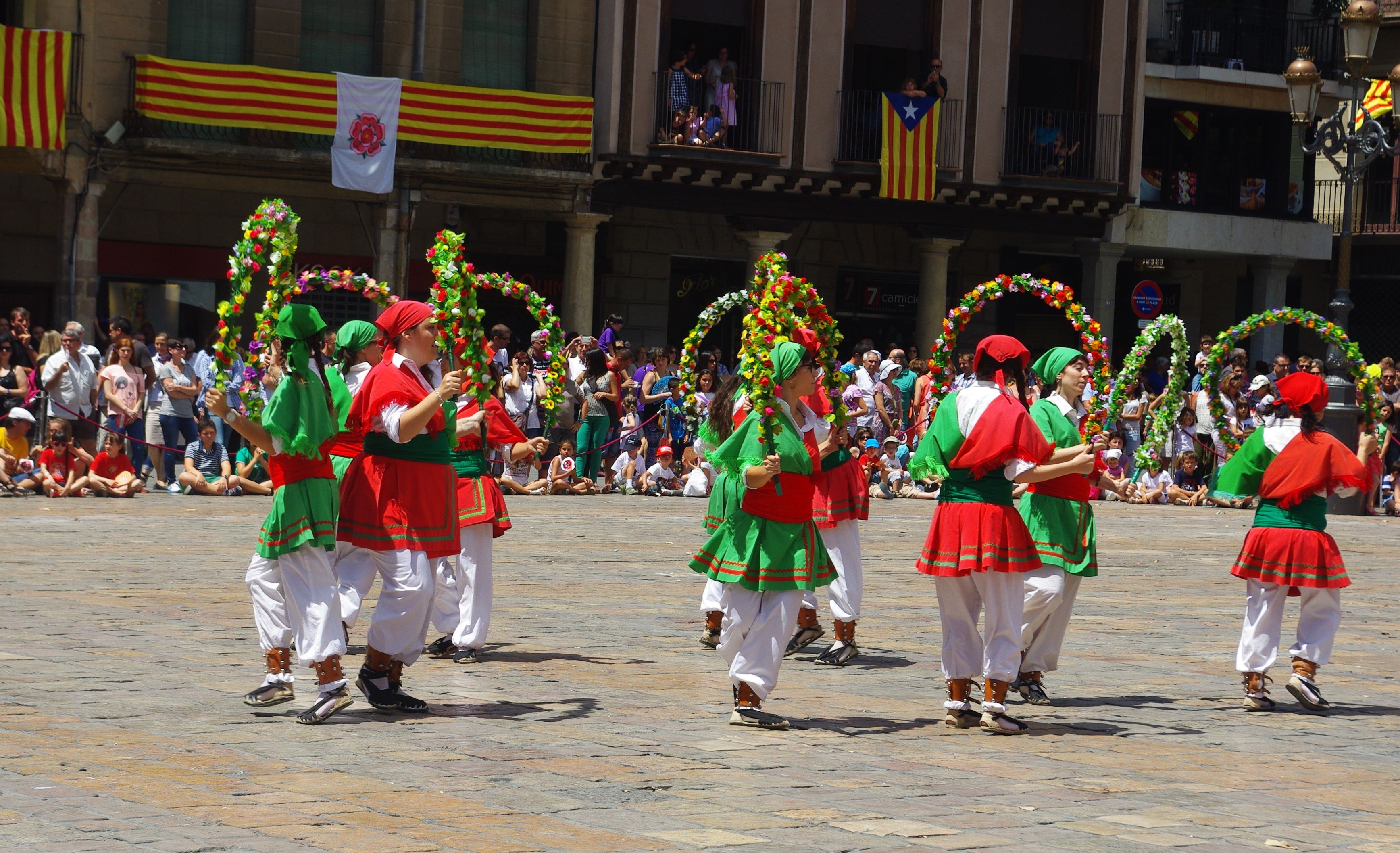 Eine Gruppe von Tänzern in grüner und roter Kleidung performs auf der Straße, umgeben von einer sitzenden und stehenden Menge, mit einem Gebäude mit Fenstern, Geländern, Säulen und Flaggen im Hintergrund.