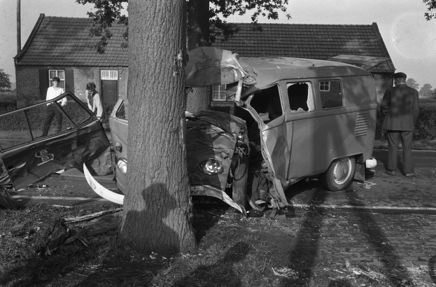 Eine schwarz-weiße Szene eines verunglückten Autos am Straßenrand mit einem Baum im Vordergrund, einigen Menschen in der Nähe und Häusern, Bäumen und einem Mast im Hintergrund.