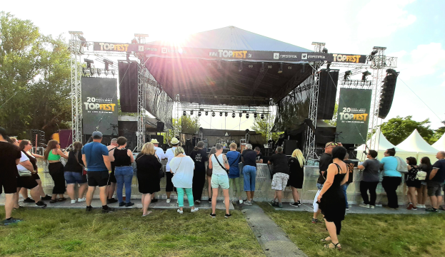 Eine Menschenmenge steht vor einer Bühne auf einem Musikfestival, mit Zelten und Bannern rechts, Gras und Bäumen im Hintergrund und einem bewölkten Himmel mit hellerer Sonne.
