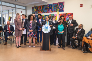 Gruppe von Menschen in einem Raum mit einem Podium, Mikrofon und einer Tafel mit der Aufschrift "Sancho-Stadtrat tritt dem Stadtrat bei", umgeben von Stühlen, Flaggen und Deckenleuchten.