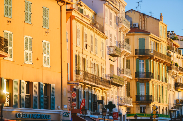 Eine Stadtstraße mit hohen Gebäuden, die Fenster, Balkone und Schilder aufweist und von Bäumen gesäumt ist, unter einem klaren blauen Himmel.