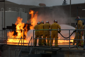 Eine Gruppe von Feuerwehrleuten in Helmen steht auf dem Dach eines Gebäudes, hält Rohre und hat Geländer, Treppen, ein Gebäude und den Himmel im Hintergrund.