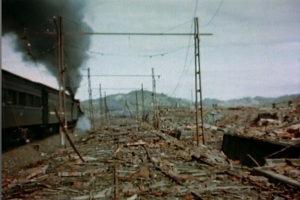 Ein Zug fährt durch ein beschädigtes Gebiet mit Rauch, der aus ihm aufsteigt, umgeben von Strommasten und -leitungen, mit Hügeln und einem klaren Himmel im Hintergrund und verstreuten Schrottteilen auf dem Boden.