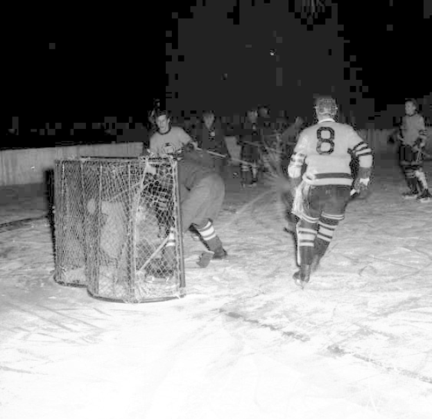 Schwarzes und weißes Foto von Männern, die Hockey auf einem Eisplatz spielen, mit einem Netz im Vordergrund und einer Wand im Hintergrund.