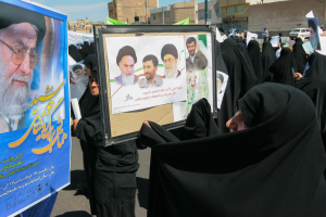 Eine Gruppe von Frauen in schwarzen Abayas hält Plakate mit Bildern von Ayatollah Ali Khamenei vor einer Kulisse aus Gebäuden, Fahrzeugen und einem klaren blauen Himmel.