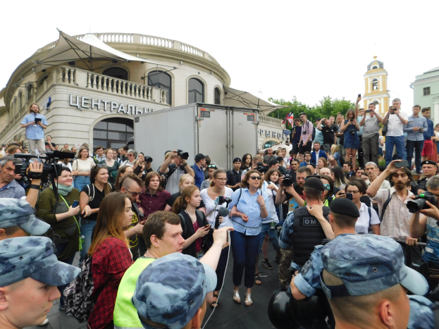Eine große Gruppe von Menschen steht vor einem Gebäude, einige halten Kameras und Telefone in der Hand, mit einem geparkten Fahrzeug, Bäumen und einem klaren blauen Himmel im Hintergrund, was auf eine Protestaktion gegen das russische Drohnenverbot hindeutet.