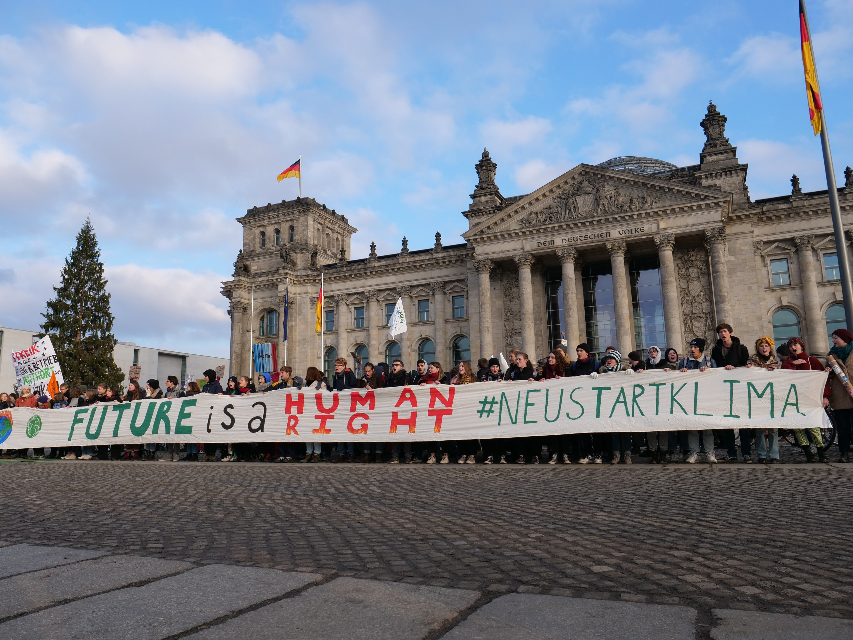 Gruppe von Menschen mit Transparent 'Zukunft ist Menschenrecht' vor dem Reichstag in Berlin, mit Säulen, Fenstern und Bögen, umgeben von Bäumen und Fahnenmasten unter einem bewölkten Himmel.