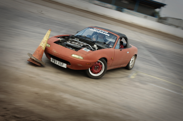 Eine Person fährt ein rotes Auto mit hoher Geschwindigkeit auf einer Rennstrecke, vor sich einen Verkehrskegel.