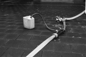 Schwarze und weiße Fotografie eines Gasbehälters mit einem Schlauch, der auf dem Boden vor einem Gebäude mit einer Tür im Hintergrund steht.