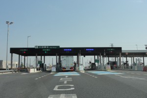 Mautstelle an der Stra├če mit einem vor ihr geparkten Lastwagen, umgeben von wei├čen und blauen Fahrbahnmarkierungen, Laternenmasten, Schildern und anderen Gegenst├Ąnden auf beiden Seiten, mit Geb├Ąuden, B├Ąumen und einem klaren blauen Himmel im Hintergrund.