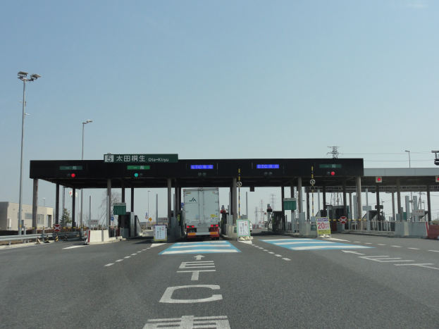 Mautstelle an der Stra├če mit einem vor ihr geparkten Lastwagen, umgeben von wei├čen und blauen Fahrbahnmarkierungen, Laternenmasten, Schildern und anderen Gegenst├Ąnden auf beiden Seiten, mit Geb├Ąuden, B├Ąumen und einem klaren blauen Himmel im Hintergrund.