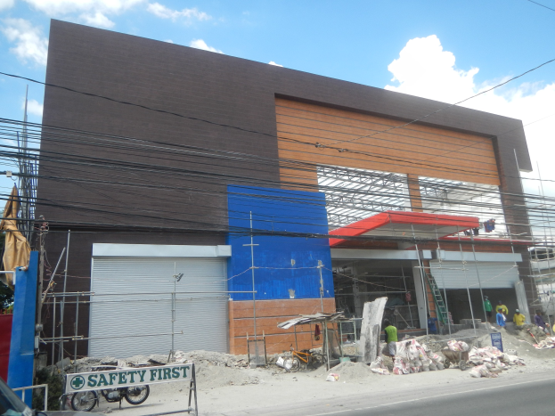 Baustelle mit einem "Sicherheit geht vor"-Schild, Fahrzeugen auf der Straße, einer Texttafel, Menschen in der Nähe, Bäumen, Strommasten, Drähten und Himmel im Hintergrund.