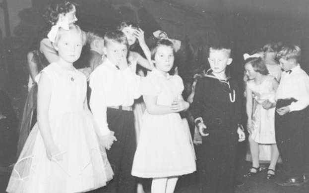 Schwarz-weiß-Foto einer Gruppe von Kindern, die auf einer Tanzfläche stehen, lächeln und glücklich aussehen, mit einer Wand im Hintergrund.