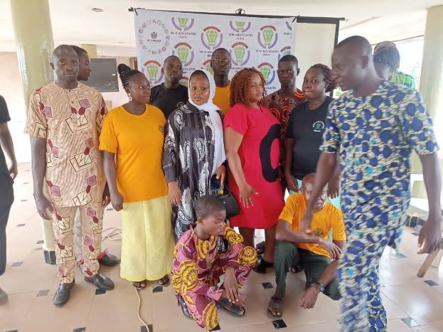 Gruppe von Menschen vor einem Banner mit der Aufschrift "Advocates in Ghana: Ein Schritt-für-Schritt-Führer" mit Stühlen, Säulen, einem Fernseher und Deckenleuchten im Hintergrund.