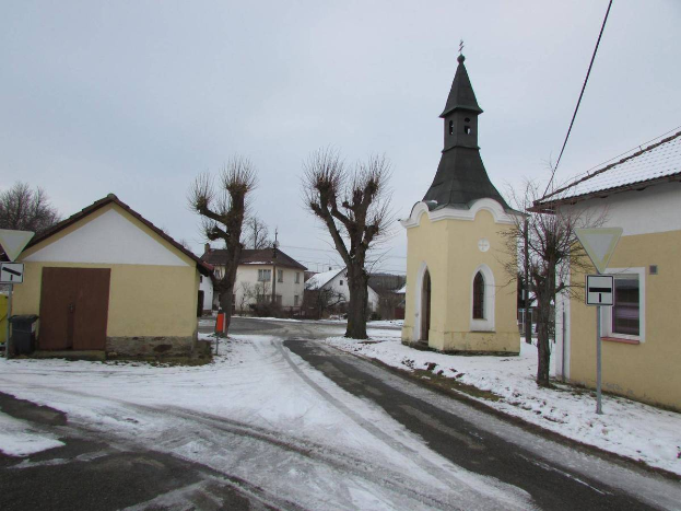 Ein kleines Dorf mit einer Kirche in der Mitte, umgeben von schneebedeckten Häusern, Bäumen und Wegweisern, unter einem bewölkten Himmel.