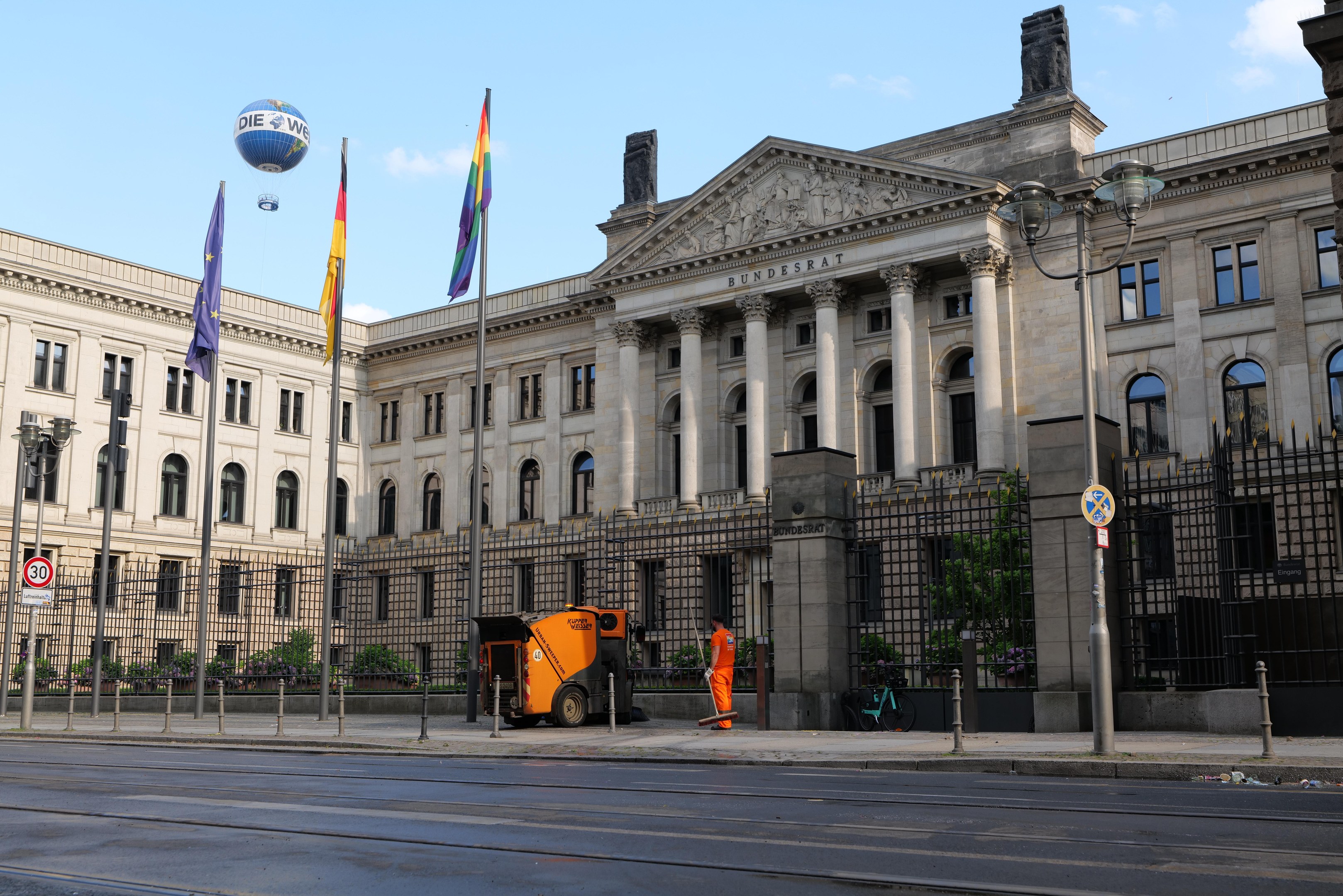 Großes modernes Gebäude mit vielen Fenstern, als Bundestag in Berlin, Deutschland identifiziert, umgeben von Straßeninfrastruktur, Fußgängern, Flaggen und Bäumen unter einem bewölkten Himmel.