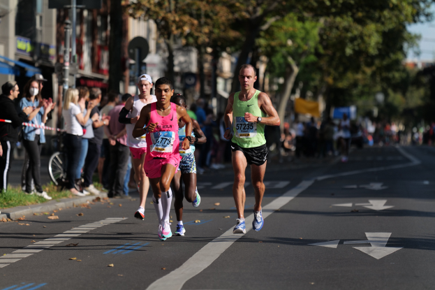 Gruppe von Läufern, die an einem Marathon auf einer Stadtstraße mit Zuschauern, Bäumen, Gebäuden und einem Fahrrad im Hintergrund teilnehmen.