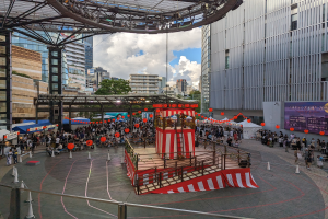 Große Menschenmenge versammelt sich auf einer Bühne in einer städtischen Umgebung zur Eröffnung des Tokyo Circus Festivals, mit Performern und Equipment im Hintergrund.