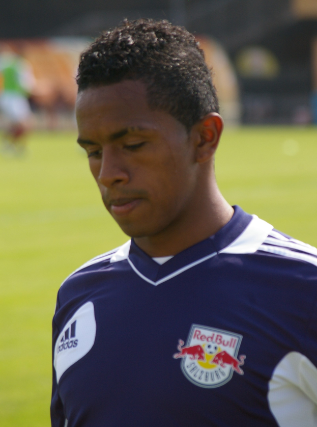 Ein junger Mann in einem blauen T-Shirt steht auf einem Fußballfeld, mit kurzen, dunklen Haaren und einem entschlossenen Gesichtsausdruck, mit einem unscharfen Hintergrund.