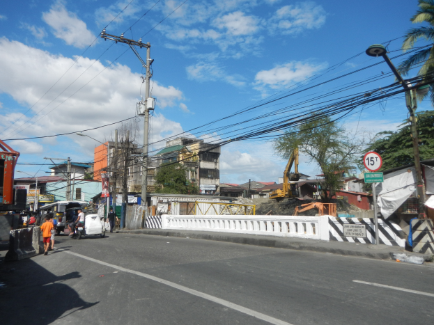 Eine Straßenszene in der Stadt mit Fahrzeugen, Fußgängern, Strommasten, Schildern, Bäumen, Gebäuden und einer Baustelle unter einem bewölkten Himmel.