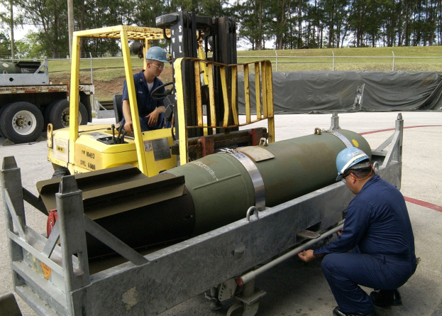Zwei Männer in blauen Uniformen und harten Helmen arbeiten an einer großen Metallfragmentierungsbombe im Freien, mit einem Zaun, Bäumen und einem klaren blauen Himmel im Hintergrund.