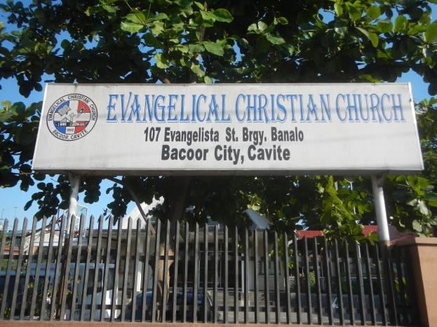 Eine evangelische Kirche in Bacoor City, Cavite, mit einem Schild, einem Metallzaun, einer Wand, Bäumen, Fahrzeugen, Gebäuden, Pfählen und einem bewölkten Himmel.