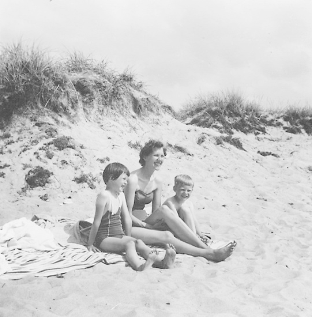 Schwarzes und weißes Foto einer Frau und zweier Kinder, die auf einem Badetuch am Strand sitzen.