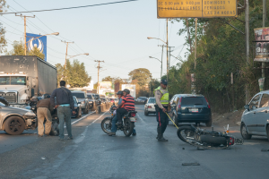 Gruppe von Menschen um ein verunglücktes Motorrad auf der Seite einer Straße mit mehreren Fahrzeugen, darunter ein Lastwagen, und Hintergrundelementen wie Bäumen, Pfählen, Lampen, Schildern und Himmel.
