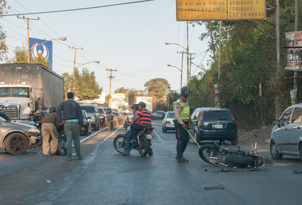 Gruppe von Menschen um ein verunglücktes Motorrad auf der Seite einer Straße mit mehreren Fahrzeugen, darunter ein Lastwagen, und Hintergrundelementen wie Bäumen, Pfählen, Lampen, Schildern und Himmel.