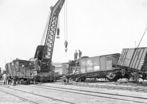Schwarzes und weißes Bild eines Zugunglücks mit einem Kran, der einen Zugwagen auf die Schienen hebt, mehreren Menschen in der Nähe und Gebäuden im Hintergrund bei klarem Himmel.