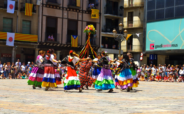 Eine Gruppe von Frauen in bunten Kleidern tanzt auf der Straße, umgeben von einer Menschenmenge, mit Gebäuden, Geländern, Balkonen, Flaggen, einem Laternenmast und einer Tafel mit Schrift im Hintergrund.