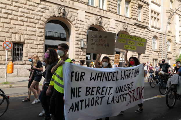 Eine Gruppe von Menschen marschiert auf einer Straße bei einer Demonstration, einige halten Schilder und andere fahren Fahrräder, im Hintergrund ist ein Gebäude mit Fenstern, Bogengängen, Säulen und Skulpturen zu sehen.