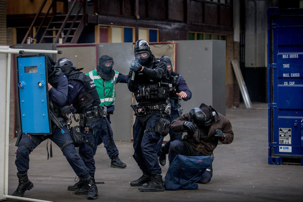 Polizeibeamte in Einsatzausrüstung stehen vor einem Gebäude in einer Reihe, einige halten Waffen, mit einer Person im Vordergrund und metallischen Gegenständen, Treppen und einer Wand im Hintergrund.