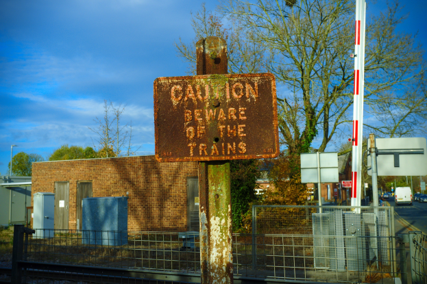 Vorsichtsschild an einem Bahnübergang-Zaun mit Bäumen, Pfählen, einem Gebäude, Containern, Fahrzeugen und einem bewölkten Himmel.