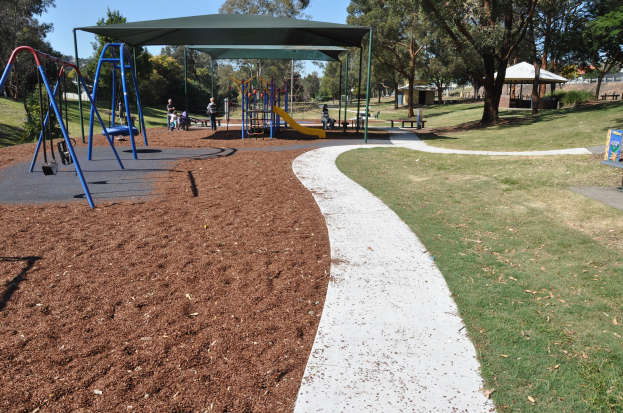 Ein Park mit einem Spielplatz, der Schaukeln, Rutschen und andere Spielgeräte, umgeben von grünem Gras, Bäumen, Bänken, Schuppen im Hintergrund und einem klaren blauen Himmel.
