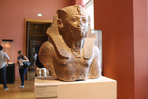 Ein Büste eines ägyptischen Pharaos in einem Museum ausgestellt mit einer Texttafel, einer Gruppe von Menschen, einer Frau mit einer Tasche, einer Tür, einer Wand und einer Decke mit Lampen.