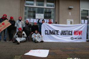 Eine Gruppe von Menschen mit Mötzen hölt ein "Climate Justice Now"-Schild vor einem gläserwändigen Gebäude mit Papieren an seiner Wand, einige Individuen sitzen auf dem Boden.